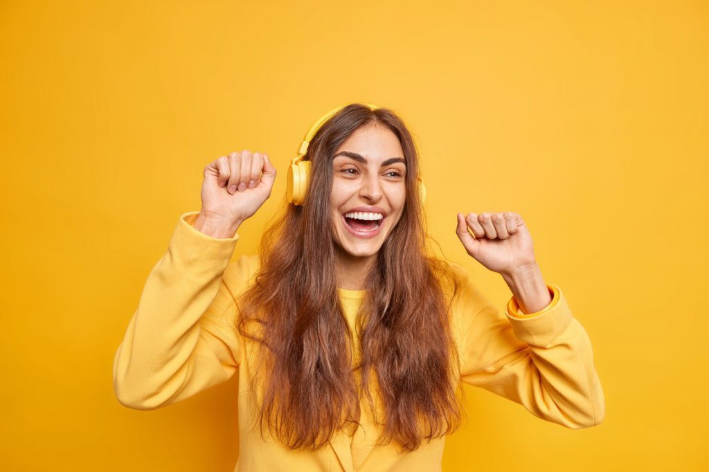 horizontal shot upbeat dark haired woman uses headphones moves body enjoys new playlist cheerful expression likes tune wears yellow jacket smiles broadly has fun 273609 54580